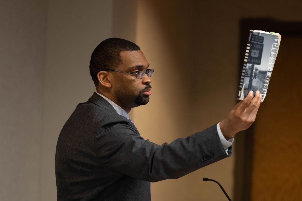 Male presenter standing at the podium holding up a book at the 2023 SOPH Research Day